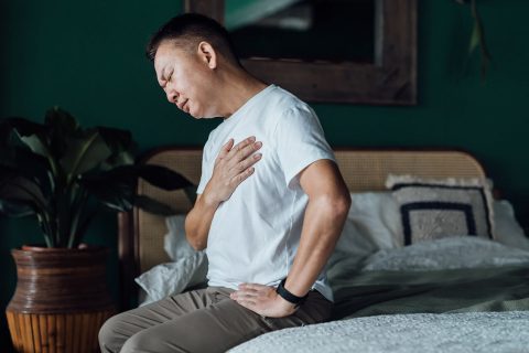 Man holding his chest in pain while sitting on a bed.